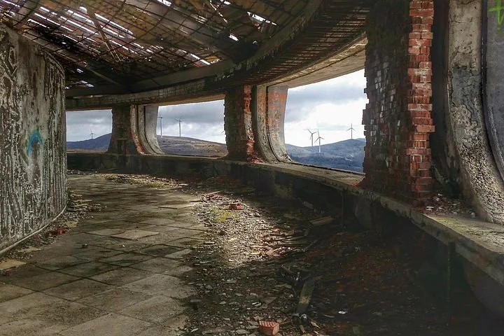 Innenansicht eines verlassenen Gebäudes mit großen Fenstern und Blick auf eine Landschaft.